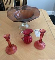 Full view of the four pieces in the lot taken on a table: two cranberry glass candlesticks, small pitcher, and the pedestal bowl