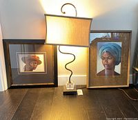 Table lamp with beige shade and two framed artworks of women on floor.