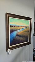 Framed signed original acrylic painting hanging on a wall, showing a river flowing through a grassy landscape underneath a soft blue sky.