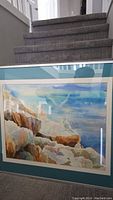 Frontal view of the framed watercolor painting showing the entire seaside scene with rocks and ocean under a bright sky.