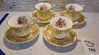 Four Paragon teacups and matching saucers arranged on a countertop, showing detailed gold filigree and floral patterns.