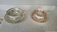 Two Paragon teacups and matching saucers with detailed gold, floral, and crown monogram decoration, photographed side by side from an angle.