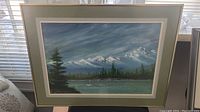 Framed original painting showing full view of snowy mountains, water with small boat, green trees and cloudy sky.