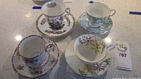 Four bone china teacup and saucer sets displayed on a countertop, showcasing different floral designs and gold trims.
