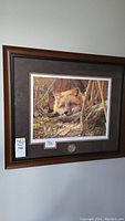 Framed art showing a fox in woods resting its head on a log with a dark brown wooden frame and matting, Ducks Unlimited medallion below print.