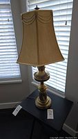 Full view of the lamp showing beige fabric shade with swagged trim, brass body, and overall height.