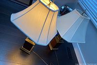 Two wood and metal table lamps with white fabric octagonal shades, one lamp lit showing warm light, both lamps resting on dark flooring near window with blinds.