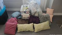 Photo shows an assortment of throw pillows in multiple colors and textures arranged on the floor, including two light green botanical pattern pillows in front, alongside duvet blanket sets mostly in plastic packaging.