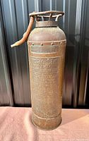 Full front view of antique copper and brass fire extinguisher showing body and hose, placed on a soft surface with dark background.