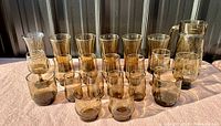 Photo showing full collection of smoky brown tinted vintage glassware, featuring multiple pitchers, drinking glasses in various sizes, and vases, all with a translucent smoky tint and rounded mid-century modern shapes.
