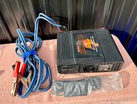 Top and front perspective of Motomaster Eliminator 3000 W power inverter laid on flat surface with jumper cables and accessory pouch beside it.