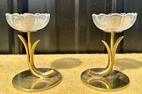 Front view of two brass and glass flower candle holders showing the curved stem, leaves, and clear glass flower cup on brass base.