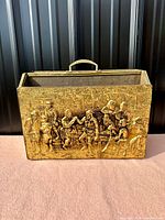 Front view of the brass relief on the wood magazine rack showing detailed scene and brass handle.