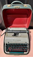 Top front view of vintage Remington Quiet-Riter typewriter in original green case with red interior lining; typewriter in green with black and white keys, showing Remington logo.