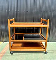 Full front view of three-tier teak bar cart on casters against metal backdrop