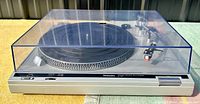 Front angle showing the Technics SL-B2 turntable with closed clear plastic dust cover on a yellow surface.