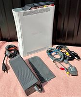 Xbox 360 console upright with power brick, cables and faceplate on pink surface with black background.