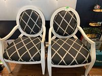Full view of the pair of white framed arm chairs with black and silver geometric lattice patterned upholstery