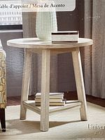 Front view of the round wooden accent table with a light finish, round top, three legs, and a lower triangular shelf with books on it.