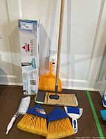Photo showing the Bissell vacuum box, orange dustpan, mop and cleaning brooms and brush on floor against wall.