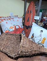 Wicker cornucopia laid flat on floor with three decorative pillows (two fall and one Halloween themed) and wooden pumpkin plaque visible in background.