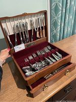 Open wooden flatware storage box showing assorted stainless steel knives, forks, and spoons with maroon velvet lining.