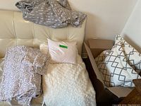 View of grey flannel mountain print fitted sheet, white textured throw blanket, pale pink pillow, white sheets with grey leafy pattern, and box of decorative pillows with grey geometric designs.