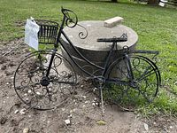 Full view of the black metal garden bicycle on grass, showing scrollwork wheels, basket and plant holding areas.