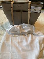 Full view of the taupe Michael Kors handbag with beige adjustable shoulder straps and protective white storage bag with gold Michael Kors lettering.