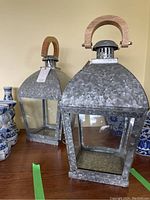 Two handcrafted metal and wood lanterns side by side, showing galvanized metal finish, clear glass panels, and wooden handles