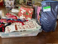 Photo showing a plastic storage bin filled with a variety of gift bags and a medium sized Christmas-themed gift box on top. Also a large blue zippered bag beside the bin labeled with lot number.