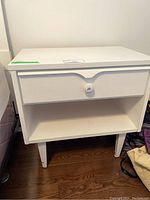 Front view of white side table with single drawer and open shelf, decorative knob visible.
