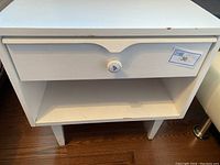 Front view of white painted mid century modern side table showing drawer and open shelf with visible wear and scratches on wood.