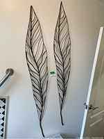 Two tall, slender metal wall decor pieces shaped like elongated leaves with detailed vein design, photographed hanging on a white wall next to a door and stair railing.