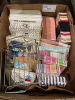 Box containing assorted new paper napkins including plain beige with printed text, striped, multicolor, and pink designs, along with a metal napkin holder placed on the napkins.