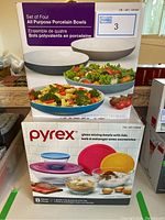 Boxes of porcelain serving bowls and Pyrex glass mixing bowls with lids stacked, showing branding and quantity information.