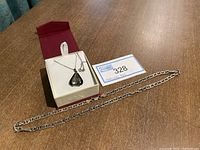 Full view of two silver necklaces on table; thick chain laid out, thin chain with pendant in box; lot card indicating Sterling 925