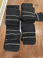 Top view of all six black bath towels and one matching washcloth arranged on wooden floor showing white dotted detail lines.