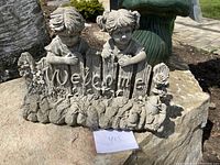Full view of concrete welcome garden sign with littl children figurines and floral decorations on a rock.