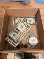 Box containing folded and flat U.S. paper currency $1 and $2 bills, assorted coins, and a collector coin in a display case.