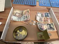Photo showing assorted paper bills from India and possibly other Asian countries along with various coins in a tin lid and scattered on the box surface including an Australian Army commemorative coin in display card