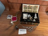 Photo showing the decorative jewelry box open with various costume jewelry including rings, brooches, watch, and necklace spread on wooden surface.