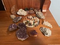 Overall group view showing amethyst geodes, rough rocks, and tumbled stones on a wooden surface with mirror base for polished stones.