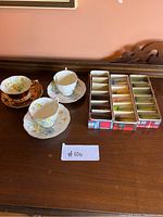 Photo showing three fine china teacups with matching saucers and boxed decorative shot glasses on display.