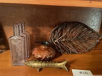 Three clear glass rectangular bottles, pottery vase with leaf pattern, metal leaf tray, and bronze fish figurine on wooden shelf.