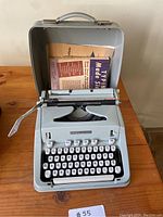 Front view of pale blue Hermes 3000 typewriter open in case, showing keyboard and instruction booklet tucked in lid.
