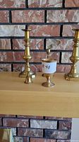Photo showing the brass items including candle holders, chalice, and bell placed on a wooden shelf against a brick wall background.