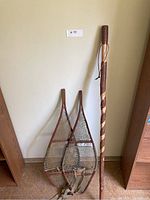Full view of two vintage wooden snowshoes and a carved wooden walking stick standing against a wall.