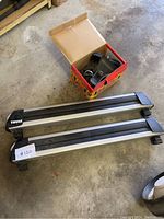 Two Thule roof rack crossbars laid on floor with a box containing mounting hardware next to them. Shows front perspective.