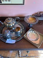 Silver plate tea set on large round tray, including teapot, sugar bowl, creamer, displayed on wooden dresser with shell-shaped bowls beside it. Tray and tea set have polished silver finish, slight tarnish visible.